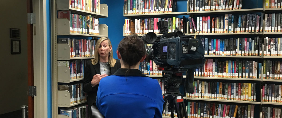 woman being interviewed in front of books, blue wall, interviewees back to viewer